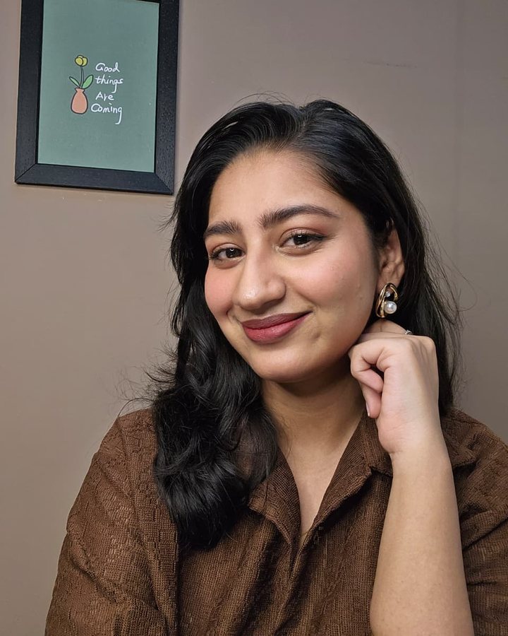 Kanchan smiling, chin resting on her hand, in a brown shirt at her filming wall